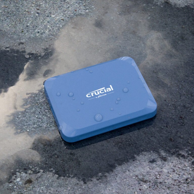 A blue Crucial X10 1TB Type-C Portable SSD with water droplets on its surface sits on wet pavement, reflecting the cloudy sky above.