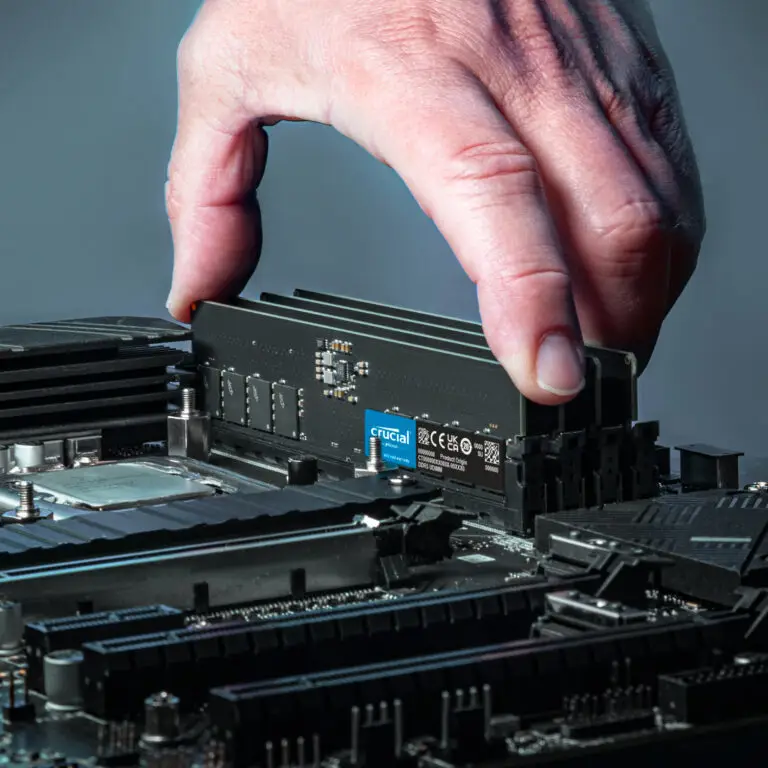 A hand installing a Crucial 32GB 5600Mhz DDR5 Desktop Memory module into a motherboard slot inside a computer, with detailed circuitry and components visible.